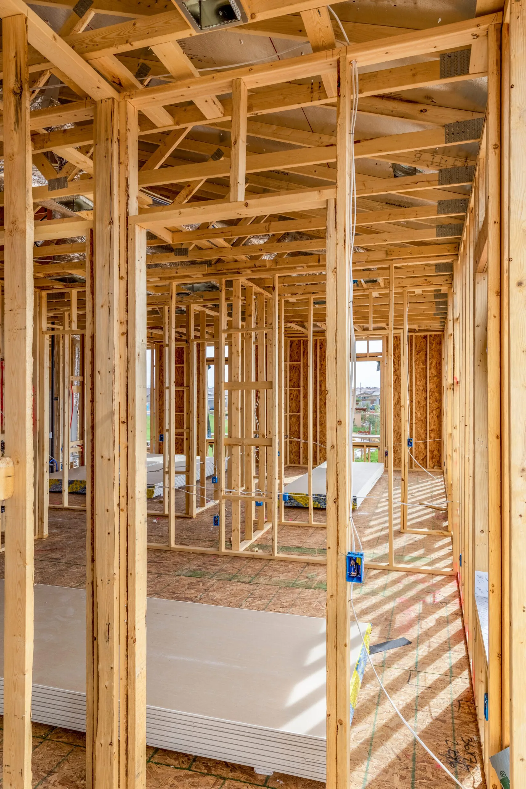 Interior view of a house under construction, showing exposed wooden beams and studs, with sunlight filtering through.
